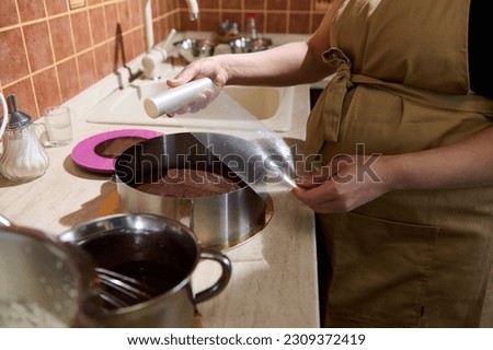 Close-up pastry chef in beige apron, rolling out a transparent border film over a kitchen countertop with food processor and ingredients, preparing for making delicious homemade cake at home kitchen