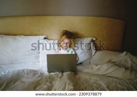 Cute adorable little toddler girl watching cartoons on laptop in bed. Tired baby child with tablet device with touch technology.