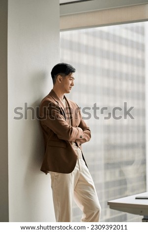 Young pensive confident Asian business man leader executive looking at big window in office. Professional male manager standing arms crossed thinking of new business ideas and goals. Vertical