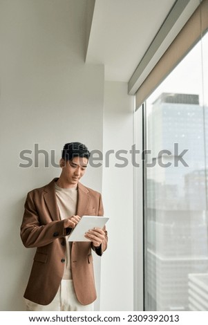 Busy smiling young adult Asian Japanese business man professional manager executive using digital tablet device fintech computer smart technology in corporate office standing at window, vertical. Royalty-Free Stock Photo #2309392013