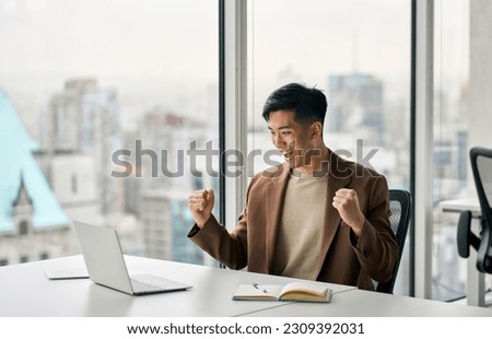 Happy young Asian business man celebrating success at work in office looking at laptop excited about financial trading growth, goals achievement good online results, screaming yes watching game. Royalty-Free Stock Photo #2309392031
