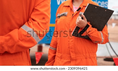 Action of a safety officer in orange coverall uniform is standing and talking with colleague during perform safety audit at construction work site. Industrial working with people photo scene.