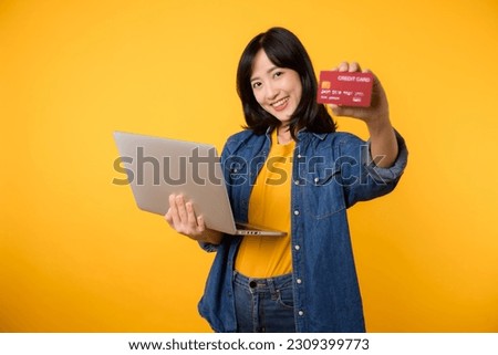 Young happy woman of Asian ethnicity wear yellow t-shirt denim shirt using laptop pc computer hold credit bank card shopping online order delivery isolated on plain yellow background.