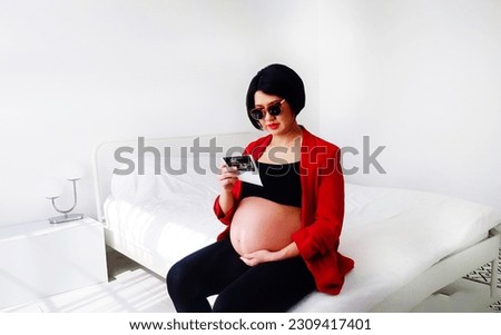 Beautiful and pretty pregnant woman wearing black glasses, sitting on bed, looking at her baby's film picture and touching her belly. Pregnancy 7-8 months, motherhood, love, expectation and care.