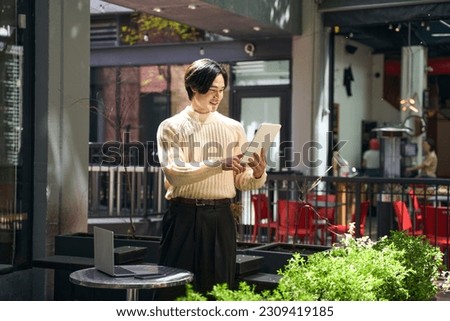Handsome smiling Japanese man wearing stylish sweater holding digital tablet looking at screen. Happy small business owner standing at the street 