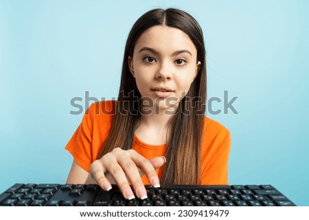 Pensive attractive teenage girl in orange t shirt typing on keyboard isolated on blue background. Concept of online education