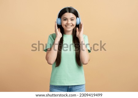 Beautiful happy latin teenage girl wearing blue headphones listening music looking at camera isolated on beige background. Modern technologies, positive lifestyle