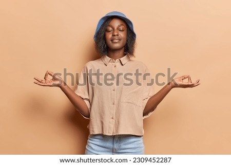 Horizontal shot of calm relaxed adult Afro woman keeps eyes closed mediatated indoor practices yoga wears panama shirt and jeans isolated over brown background. Keep calm. Relaxation exercise