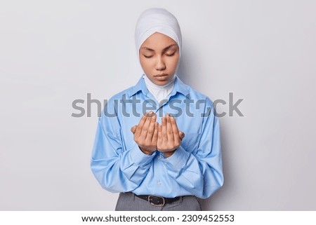 Muslim woman makes traditional prayer to God wears hijab and blue shirt keeps eyes closed prays indoor against white background. Islamic female model prays. Praying concept. Meditation concept