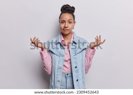 Latin woman raises her shoulders in shrug has puzzled expression as if searching for answers expresses confusion and uncertainty feels indecisive wears denim clothes isolated over white background