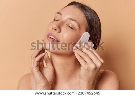 Skin treatment and beauty routine concept. Pleased brunette European woman massages face with jade stone touches cheek gently has eyes closed isolated over brown background enjoys sensation.