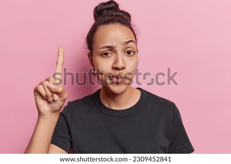 Selfassured and confident woman points index finger upward demonstrates something important raises eyebrows dressed in casual black t shirt isolated over pink background. You should check this out Royalty-Free Stock Photo #2309452841