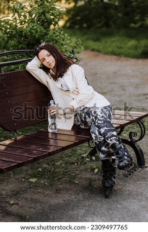 Beautiful smiling woman, girl athlete sits on a wooden bench in the park in spring in roller skates with a bottle of water, resting after a ride. Photography, portrait, sports, recreation.