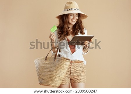 Beach vacation. smiling trendy housewife in white blouse and shorts on beige background with credit card, straw bag and summer hat using tablet PC. Royalty-Free Stock Photo #2309500247