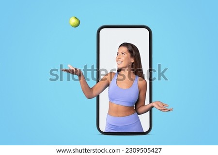 Glad young european slim lady athlete brunette in sportswear tossing green apple on screen of big smartphone, isolated on blue studio background. Sports blog, fit, app for body care and vitamins