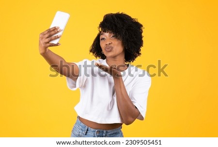 Happy young african american curly lady make blowing kiss and selfie on smartphone, have fun, isolated on yellow studio background. Social networks, beauty care, blog and photo app