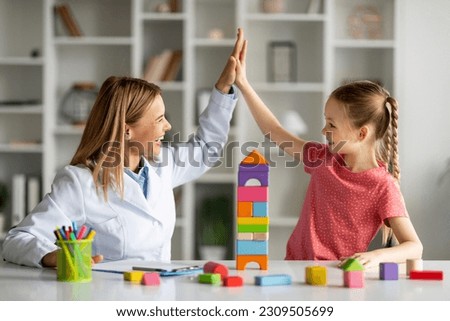 Cute Little Girl Giving High Five To Child Development Specialist During Meeting In Office, Smiling Therapist Lady Greeting Happy Female Kid With Progress, Celebrating Successful Therapy Royalty-Free Stock Photo #2309505699