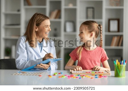 Happy Child Development Specialist Exercising With Little Girl And Taking Notes, Cheerful Female Kid Making Word With Colorful Letters On Table, Enjoying Lessons With Logopedist Lady, Closeup Royalty-Free Stock Photo #2309505721