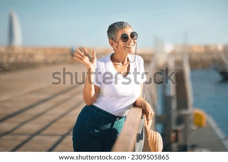 Hello, Golden Summer. Smiling Mature Woman Waving Hand Posing At Marina Pier With Sailboats Outdoor, Meeting Her Retirement Life And Holidays By The Sea. Happy Travel Lifestyle Royalty-Free Stock Photo #2309505865