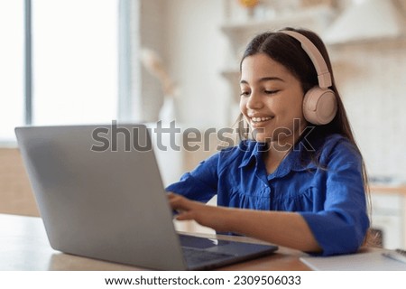 E-Learning Class. Cheerful Arabic Schoolgirl Studying Online Typing On Laptop Computer Wearing Earphones Sitting At Home. School Kid Girl Websurfing Enjoying Distance Lesson Royalty-Free Stock Photo #2309506033