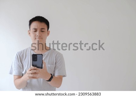 A young Asian man who wears a grey t-shirt and a smartwatch is holding and looking at the smartphone with confused