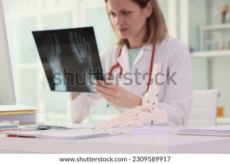Woman doctor in work uniform examines bone injury on X-ray next to human foot model sitting at workplace in modern clinic office. Bone fracture concept