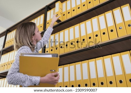 Serious female clerk takes folders from rack in large archive premise. Accountant checkups old documents in office storage. Paperwork at company Royalty-Free Stock Photo #2309590007
