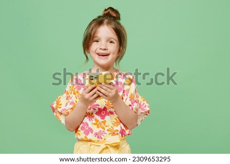 Little cute child kid girl 6-7 years old wear casual clothes hold in hand use mobile cell phone isolated on plain pastel green background studio portrait. Mother's Day love family lifestyle concept