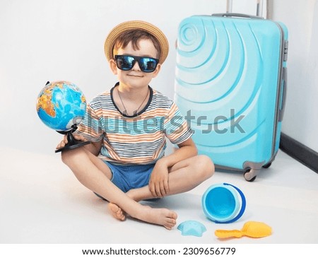 A cute boy in a hat and sunglasses in bright clothes sits on a light background near a blue suitcase with a globe in anticipation of a trip and adventure