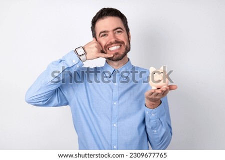 businessman wearing blue shirt holding a piggy bank  over white background smiling doing phone gesture with hand and fingers like talking on the telephone. Communicating concepts.