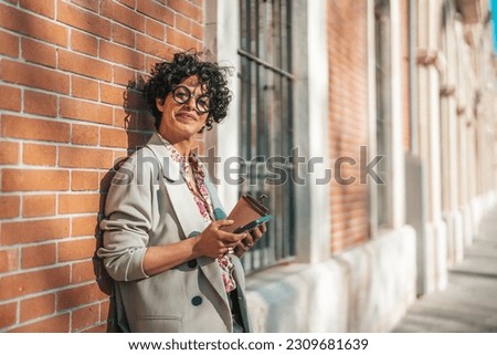 Lifestyle portrait of a mature and cheerful woman standing with phone and coffee cup on the public transport stop outdoors. Urban business travel and transportation concept. 