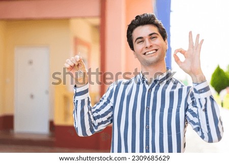 Young handsome man holding home keys at outdoors showing ok sign with fingers