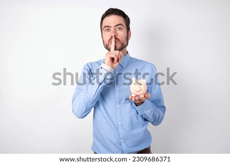 businessman wearing blue shirt holding a piggy bank  over white background makes silence gesture, keeps finger over lips. Silence and secret concept.