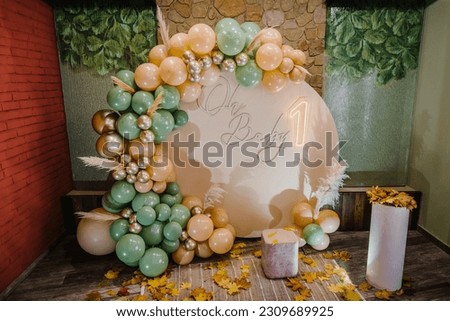Arch decorated green, brown, golden balloons, text oh baby, neon number one, dry leaves. Photo wall decoration trendy autumn decor. Birthday party for 1 year old girl or boy. Children's photo zone. Royalty-Free Stock Photo #2309689925