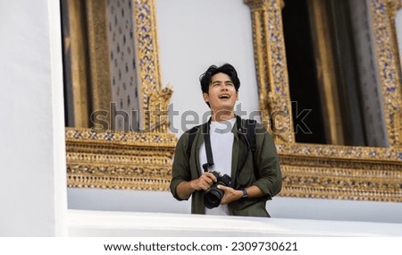 Portrait of Asian male travel photographer holding a camera in front of buddhist temple on street in Bangkok, Thailand, Southeast Asia. Handsome man taking a picture while traveling.
