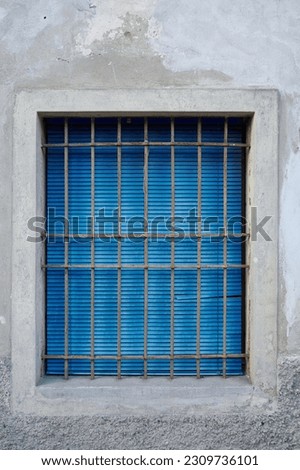 The window of the old house is closed by a worn blue Venetian blind, which shows the signs of the past. A sturdy iron grate protects it