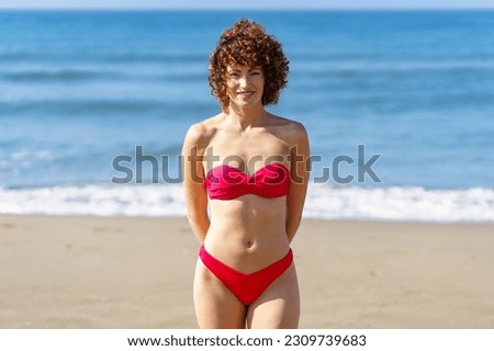 Delighted curly haired female in red swimwear standing on sandy shore against rippling sea and looking at camera in sunlight
