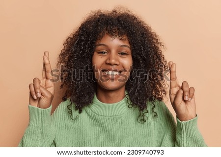 Positive African woman smiles toothily keeps fingers crossed believes in good luck makes wish anticipates results dressed in green knitted jumper isolated over brown background. Body language concept