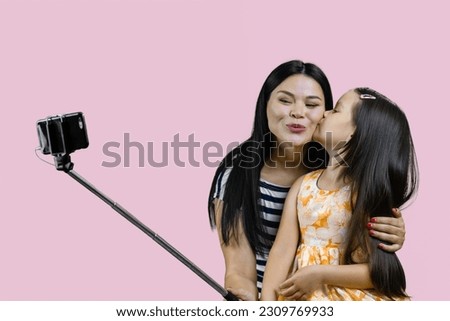 Mother and her daughter are making a selfie on smartphone using the stick. Little girl kissing her mom for photo. Isolated on pink.