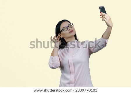 Cute young asian woman in glasses is taking a selfie on her smartphone. Isolated on yellow.