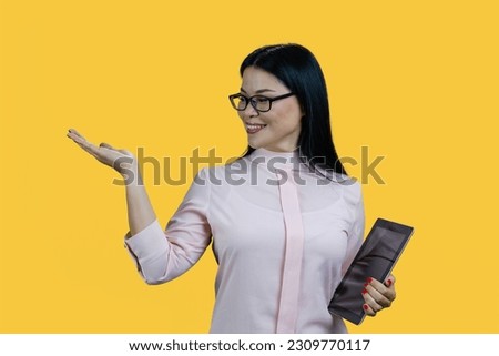 Happy asian woman in glasses is holding tablet pc. Advertising copy space with palm up. Isolated on yellow background.