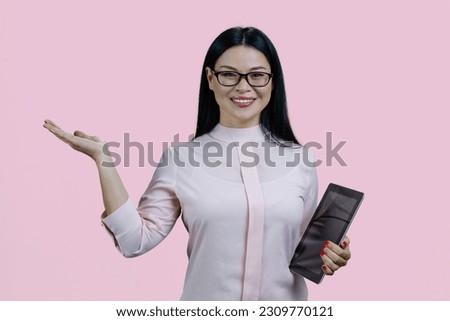 Young smart asian woman in glasses is holding tablet pc and advertising copy space. Isolated over pink background.