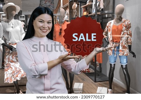 Smiling brunette asian woman advertising clothing shop discount. Boutique in the background.