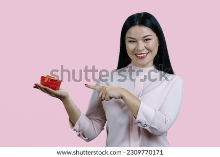 Portrait of a young asian woman is pointing at red gift box. Isolated on pink background.