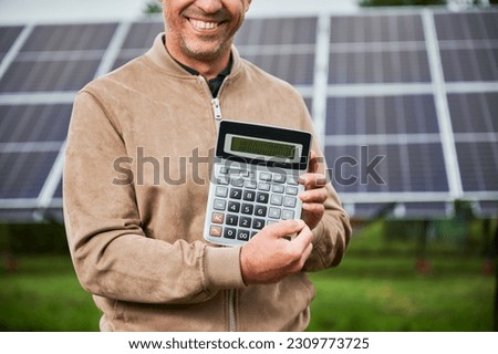 Smiling man showing future income from investing in alternative energy. Cropped picture of happy man demonstraiting big number on calculator. Man glad about huge sum profit. Royalty-Free Stock Photo #2309773725