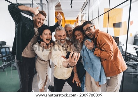 Group of laughing diverse friends hugging each other while looking at screen with excitement and standing near glass wall in office while taking self shot on smartphone Royalty-Free Stock Photo #2309780399