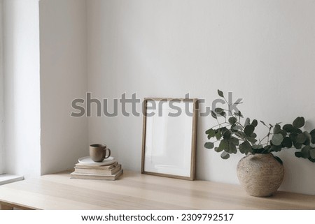 Cup of coffee, books. Empty vertical picture frame mockup on wooden desk, table. Vase with silver eucalyptus tree branches. Minimal working space, home office. Scandinavian interior design. Side view.
