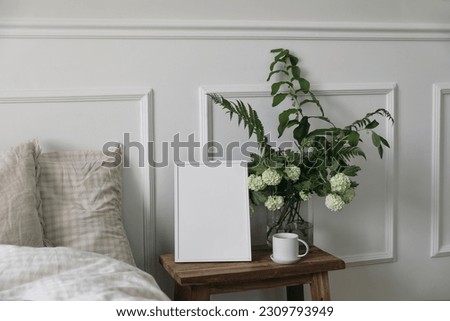 White picture frame mockup and cup of coffee on wooden night stand. White viburnum, fern and solomons seal flowers bouquet. Bedroom view. Beige pillows, linen blanket in bed. Home interior decor.