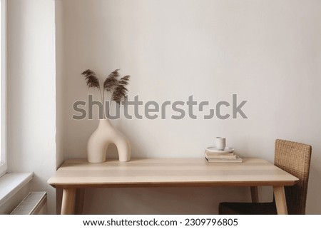 Wooden table, desk near window. Modern organic shaped vase with dry flowers, grass. Cup of tea, coffee, old books. Rattan chair at home. Minimal Scandi boho interior. Beige wall background mockup.