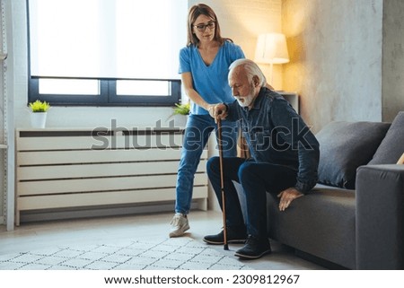 Smiling senior man with female healthcare worker. Home carer supporting old man to stand up from the chair at care home.. Female caregiver helping senior man get up from couch in living room - Indoors Royalty-Free Stock Photo #2309812967